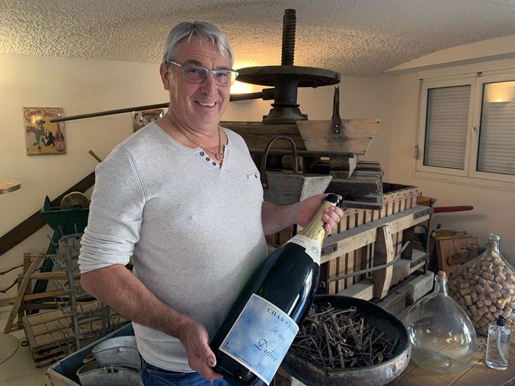 Portrait de Didier Lapie, qui tient une bouteille de champagne, devant un ancien pressoir