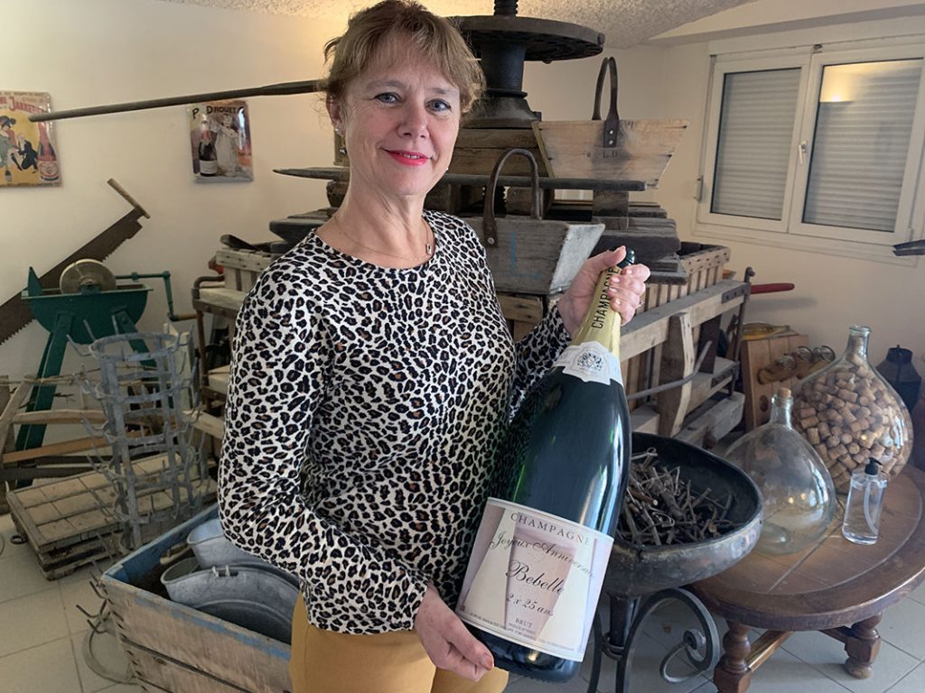 Portrait de Isabelle Lapie, qui tient une bouteille de champagne, devant un ancien pressoir