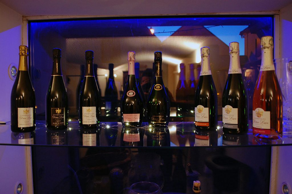 Photographie, intérieur. L'ensemble des cuvées Champagne Didier Lapie : toutes les bouteilles sur une étagère en verre
