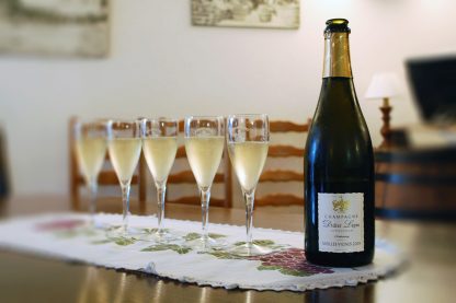 Photographie, intérieur. Une bouteille de champagne est débouchée, et à côté sont servies 5 flûtes de champagne. Texte étiquette de la bouteille : Vieilles vignes 2009